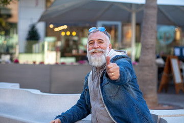 Older man with white beard and sunglasses sitting on a white bench resting with his finger up