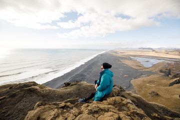 male traveler in green clothes and  sitting near a cliff view of a black beach and the Atlantic Ocean in Iceland