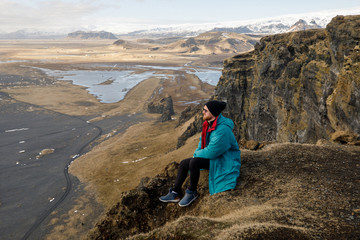 Fototapeta premium male traveler in green clothes and sitting near a cliff view of a black beach and the Atlantic Ocean in Iceland