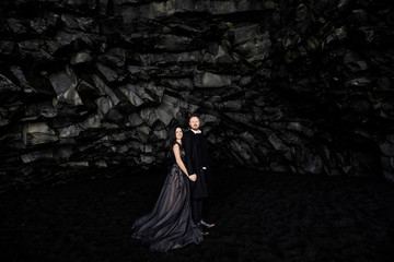 Fototapeta premium Young beautiful couple bride and groom in black clothes walks in a black cave on black sand beach near the Atlantic Ocean in Iceland