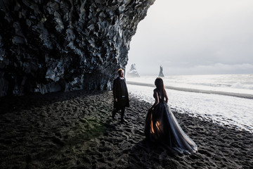 Young beautiful couple bride and groom in black clothes walks in a black cave on black sand beach near the Atlantic Ocean in Iceland
