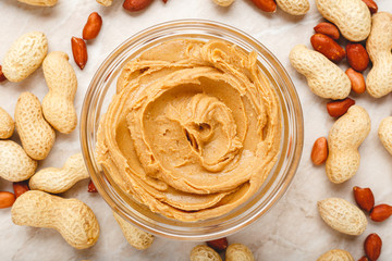 Creamy peanut paste, peanut butter in open glass jar in the center of peanuts food background. Peanuts in shell, peeled peanuts on white background . Vegan food concept. Creamy peanut paste flat lay.