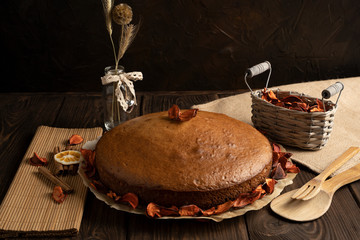 Homemade cake decorated with dried flower petals on parchment paper, next to a straw napkin, cinnamon sticks, lemon, basket and wooden appliances on a natural wooden background.