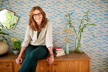 Mature woman smiling while relaxing on a cabinet at home