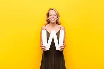 young blonde woman excited, happy, joyful, holding the letter W of the alphabet to form a word or a...
