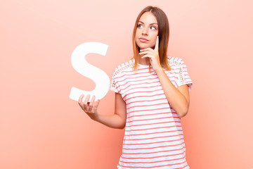 young pretty red head woman confused, doubtful, thinking, holding the letter S of the alphabet to...