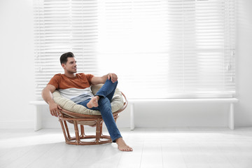 Attractive man relaxing in papasan chair near window at home. Space for text