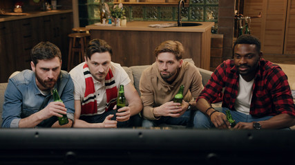 Multi-ethnic group of young flat mates watching sports competition on TV at home. They cheering up, raising hands and yelling while supporting favorite team. Friendship goals, having fun, emotions