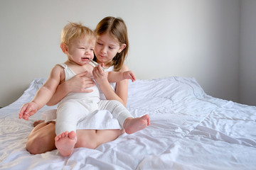 brother and older sister dressed in white clothes. A little boy sit on the lap of an older girl and she measures the temperature with a thermometer