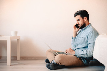 Man talking on smart phone using laptop sitting on floor against wall