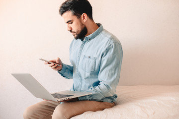 Man holding laptop and smart phone while sitting on bed at home