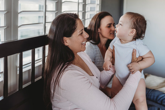 Two Smiling Moms Holding Baby Daughter At Home Near Window