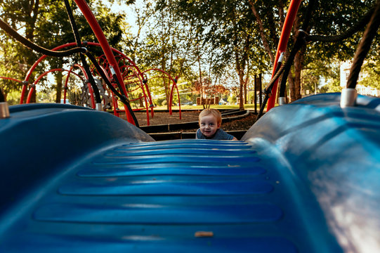 Baby boy climbing blue playground equipment at park with trees.