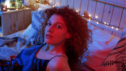 Above view of pretty female in peignoir lying on bed and looking at camera with seductive smile in pink neon light. Romantic atmosphere, seduction, dating. Playful mood. Female portrait, close up view