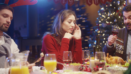 Unhappy pretty woman in red sweater feeling alone on Christmas party. Happy bearded guys talking and enjoying food at dining table.