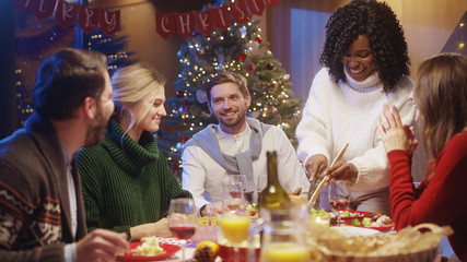 On Christmas Eve, joyful company of best friends eating delicious homemade meals. Happy afro-american girl hostess carving hot turkey talking with people.