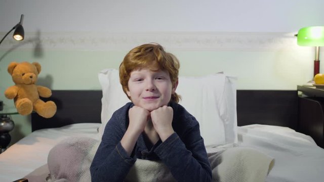 Headshot of cute Caucasian little boy switching channels, making interested facial expression, and watching TV. Redhead child looking for interesting cartoons. Childhood, leisure, bedtime.