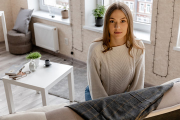 cute young girl in a white sweater and jeans sits on a sofa and looks at the camera