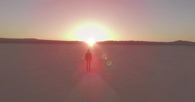 Guy wearing torn jeans and a hat stands in the lowland with the sun rising in the background.