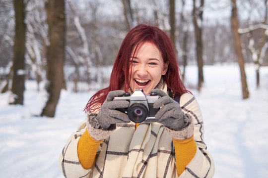 Young redhead smiling female in warm clothes with retro film camera shooting a photo on the background of winter forest, snow 