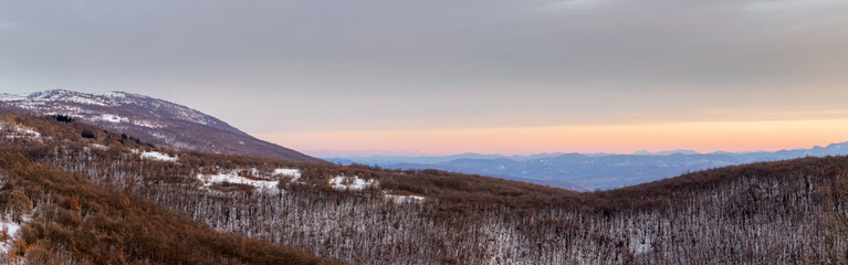 Obraz na płótnie Canvas Panoramic view of a forest on a snow covered mountain with distant mountain layers and soft colors of a sunset sky