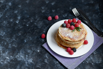 A stack of delicious pancakes with raspberries, blackberries and blueberries. On a dark stone background. Sprinkled with icing sugar and decorated