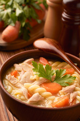 Close up of a bowl of chicken noodle soup with a wooden spoon in and hand mills with carrots and parsley in the background