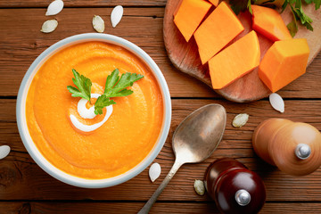 Top view pumpkin soup in bowl in and raw pumpkin on cutting board on wooden background