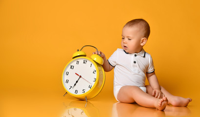 Baby boy toddler is occupied with sitting at big yellow alarm clock with hands playing showing on yellow with copy space