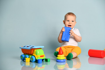Smiling infant baby boy toddler in yellow pants is sitting surrounded by toys, is holding blue...