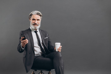 Business and fashionable bearded mature man in a gray suit sitting and using smartphone on the grey background.