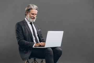 Business and fashionable bearded mature man in a gray suit smiling and using laptop on the grey background.