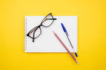 School and office supplies flay lay on colorful yellow background. Glasses, sketchbook, pencil, pen...