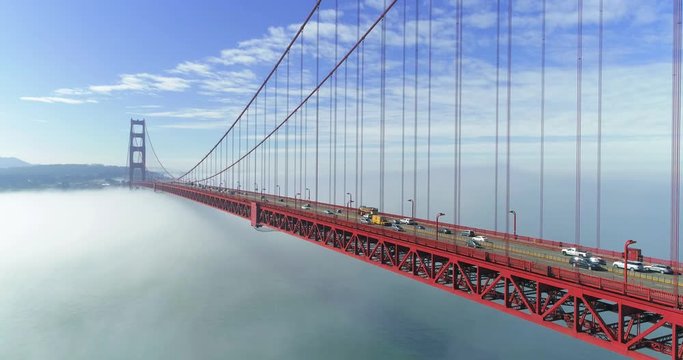 Aerial view along the deck of the Golden Gate Bridge in San Francisco, California. Cars drive between the tall red steel cables in a moody atmospheric scene while low fog hides the bay below.