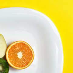 Top view of the sliced orange apple and kiwi friut at the white dish lies on the bright yellow...