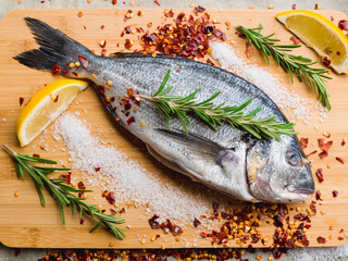 Fresh uncooked dorado or sea bream fish with lemon slices, spices, herbs and rosemary on cutting board. Mediterranean cuisine. Top view. Healthy and diet food concept. Raw fish for cooking