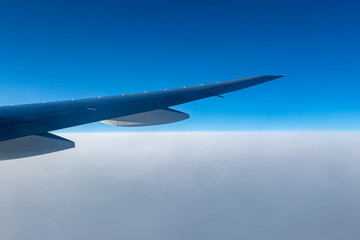 Looking out of an aircraft window at blue sky above smooth white clouds, with the aircraft wing visible