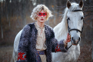 A young attractive man with curly blond hair with an ethnic ritual pattern on his face and ethnic clothing of Eastern European Slavs keeps a light-suited horse under the knot.