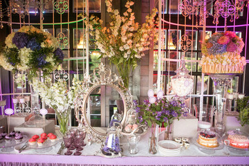 A sweet table with desserts and sweets in purple colors. Cake in pink colors on a glass stand, cupcakes, marshmallows surrounded by flowers. Decor luxury wedding celebration.