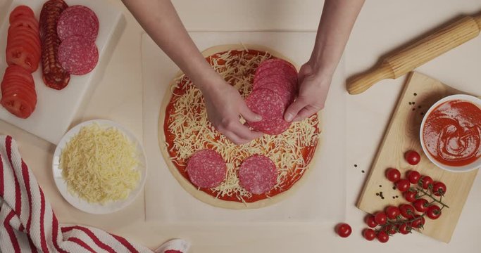 Top view of restaurant chef putting salami slices on pizza dough with tomato sauce and grated cheese on it