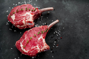 two raw steaks, beef tomahawk with spices, thyme on a stone background. with copy space for your...