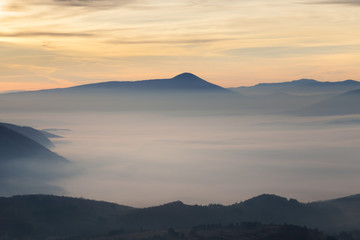 Valley filled with soft mist, distant silhouettes of impressive mountains and amazing colors of vivid morning sky