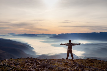Mountain hiker standing at the edge of mountain peak with hands in the air and looking at a valley filled with thick fog during colorful sunrise