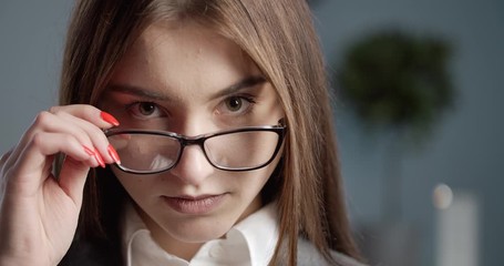 Portrait of attractive young woman with brown eyes looking at camera and adjusting eyeglasses. Professional businesswoman in formal wear standing at modern office.