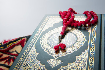 Selective focus of Holy Quran with arabic calligraphy meaning of Al Quran and red tasbih or rosary beads over white background.