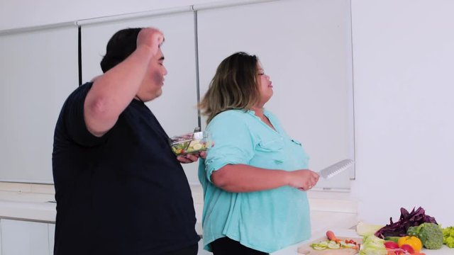 Cheerful overweight couple making vegetable salad while dancing in the kitchen at home. Shot in 4k resolution