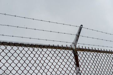 Barbed wire and chain link fence against a grey sky. The fence has three strands of barbed wire on...