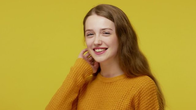 Beautiful good-natured girl in pullover with kind face expression and perfect smile looking playfully, winking at camera and adjusting her hair, flirting. studio shot isolated on yellow background