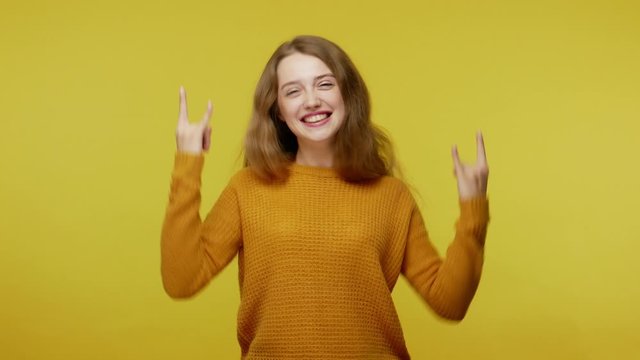 Disobedient cute girl with brown hair in pullover making rock and roll hand gesture, cool sign and showing tongue, feeling crazy carefree, naughty teenager. studio shot isolated on yellow background