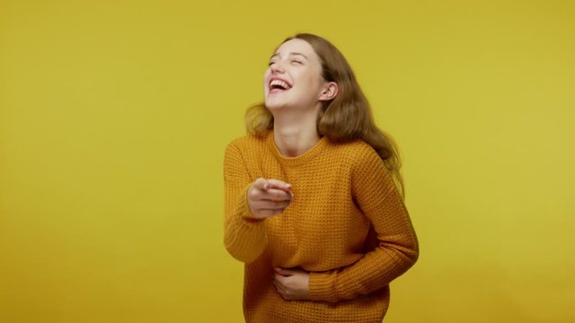 Amused happy girl with brown hair in pullover holding her stomach while laughing hysterically and pointing to camera, taunting, making fun of you. indoor studio shot isolated on yellow background